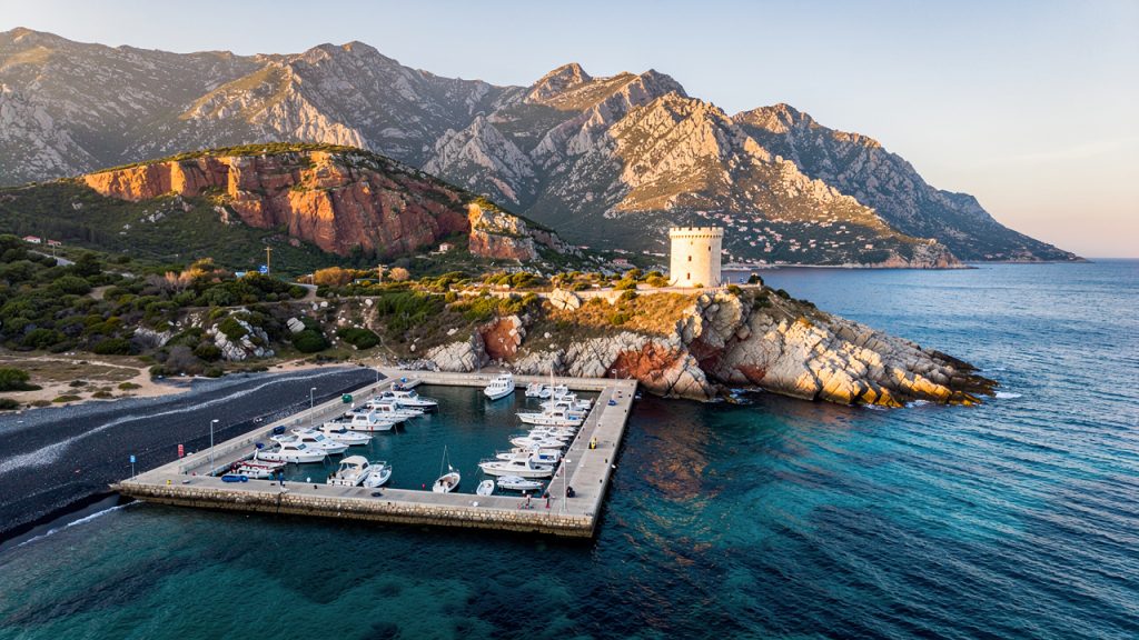 Vue aérienne réaliste de la marine de Porto en Corse, avec une plage sombre, une tour génoise, la mer Méditerranée et des montagnes escarpées baignées par une douce lumière matinale.